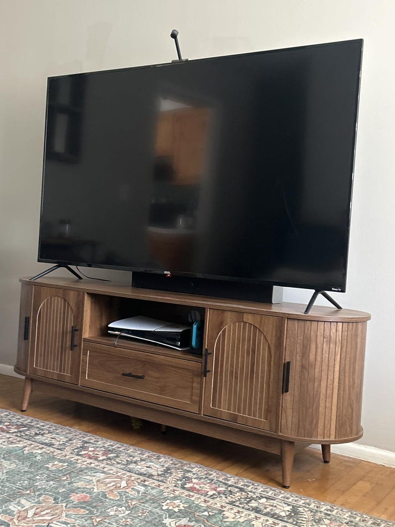 Pode incluir: Um m&oacute;vel de TV de madeira com um design moderno de meados do s&eacute;culo. O m&oacute;vel tem duas portas com ranhuras verticais, uma gaveta e uma prateleira. Uma grande televis&atilde;o preta est&aacute; em cima. O m&oacute;vel &eacute; castanho com ferragens pretas.