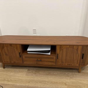 Pode incluir: Um suporte de TV de madeira marrom com um design moderno de meados do s&eacute;culo. O suporte tem duas portas com ranhuras verticais, uma gaveta e uma prateleira aberta. O suporte est&aacute; sobre pernas de madeira e tem um tampo arredondado.