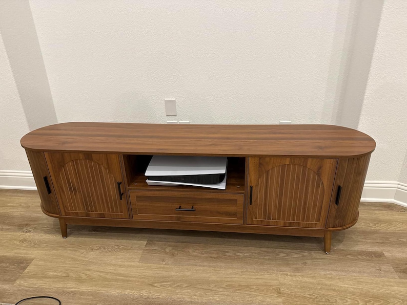 Pode incluir: Um suporte de TV de madeira marrom com um design moderno de meados do s&eacute;culo. O suporte tem duas portas com ranhuras verticais, uma gaveta e uma prateleira aberta. O suporte est&aacute; sobre pernas de madeira e tem um tampo arredondado.