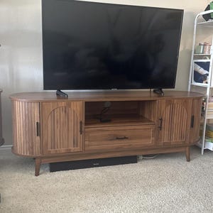 Pode incluir: Um m&oacute;vel de TV em estilo moderno de meados do s&eacute;culo, com acabamento em castanho quente. O m&oacute;vel apresenta duas portas de arm&aacute;rio com detalhes de ranhuras verticais, uma gaveta central e prateleiras abertas. O m&oacute;vel &eacute; suportado por pernas afuniladas e tem uma barra de som preta por baixo.
