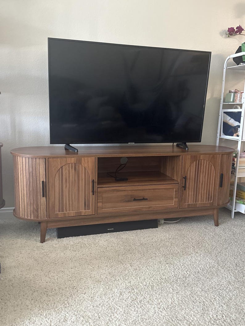 Pode incluir: Um m&oacute;vel de TV em estilo moderno de meados do s&eacute;culo, com acabamento em castanho quente. O m&oacute;vel apresenta duas portas de arm&aacute;rio com detalhes de ranhuras verticais, uma gaveta central e prateleiras abertas. O m&oacute;vel &eacute; suportado por pernas afuniladas e tem uma barra de som preta por baixo.