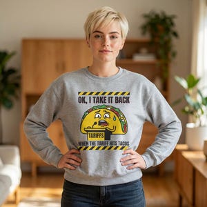 May include: A grey sweatshirt featuring a cartoon taco with a distressed expression. The text on the sweatshirt reads "OK, I TAKE IT BACK" and "TARIFFS WHEN THE TARIFF HITS TACOS". The sweatshirt is worn by a person.