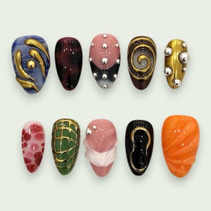 Op de afbeelding: Een set van tien kunstnagels met diverse ontwerpen, waaronder blauwe en gouden wervelingen, donkerrood schildpadpatroon, roze met zilveren studs, gouden spiralen, metallic goud met zilveren bolletjes, rode stippen, groene rasterpatronen, roze golven, zwart met goud en oranje ribbels.