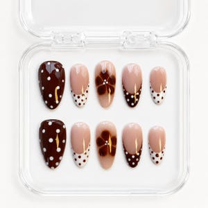 Op de afbeelding: Een set van tien ovale valse nagels in een doorzichtige plastic doos. De nagels hebben verschillende ontwerpen, waaronder donkerbruin met witte stippen, nude met een witte stippen tip en nude met een bruin bloemmotief.