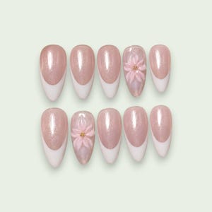 Può includere: Un set di dieci unghie finte a mandorla con una base rosa chiaro scintillante e punte bianche alla francese. Due delle unghie presentano un design floreale tridimensionale in rilievo color rosa chiaro al centro, arricchito da minuscole perline dorate.