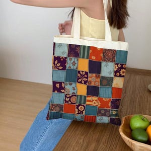 Op de afbeelding: Een patchwork draagtas met crèmekleurige handvatten. De voorkant van de tas heeft een kleurrijk patchworkontwerp met bloemen- en geometrische patronen in tinten blauw, oranje, paars en goud. De tas wordt vastgehouden door een persoon die een gele top en een blauwe spijkerbroek draagt.