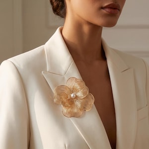 Pode incluir: Um broche de metal dourado em forma de flor com pétalas delicadas semelhantes a fios e uma única pérola branca no centro, preso na lapela de um blazer creme. O design floral apresenta linhas metálicas finas e complexas.