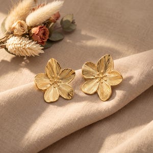Könnte beinhalten: Ein Paar goldfarbene Metall-Ohrstecker in Form von fünfblättrigen Blumen. Jeder Ohrring weist ein texturiertes, strahlenförmiges Linienmuster auf den Blütenblättern und ein kleines, erhabenes Detail in der Mitte auf. Polierte Metalloberfläche.