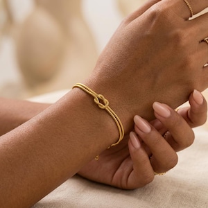 Può includere: Un braccialetto dorato con un design a nodo, indossato al polso. L'immagine mostra il design del braccialetto, con altri anelli d'oro sulle dita. Lo sfondo neutro mette in risalto l'eleganza del gioiello.
