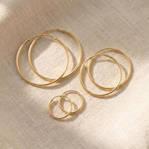 Könnte beinhalten: Eine Sammlung goldfarbener Creolen in verschiedenen Größen. Die Ohrringe sind auf einem neutralen Stoff angeordnet, wobei das Licht weiche Schatten erzeugt. Die Reifen reichen von klein bis groß und zeigen ein minimalistisches Schmuckdesign.