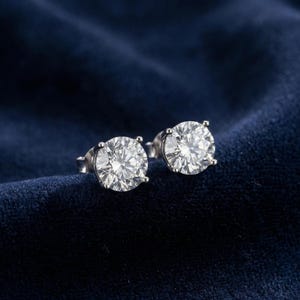 Könnte beinhalten: Ein Paar funkelnde Diamant-Ohrstecker. Jeder Ohrring hat einen großen, runden Diamanten im Brillantschliff, der in eine Silber- oder Weißgoldeinfassung gefasst ist. Die Ohrringe werden auf einem dunkelblauen Samthintergrund präsentiert.