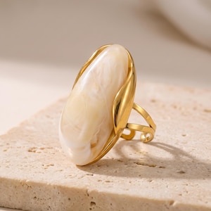 May include: A gold-toned ring featuring a large, elongated, oval-shaped cream-coloured stone with a marbled texture. The metal setting has a smooth, polished finish that curves around the sides of the stone, and the band is open and adjustable.