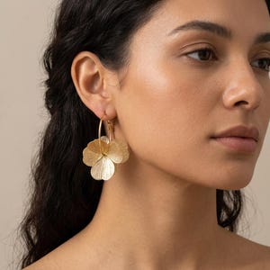 Puede incluir: Pendientes de aro dorados con diseño floral. Los pendientes presentan tres pétalos de flores texturizados, creando un efecto en capas. Los aros son finos y circulares, y las flores están unidas en la parte inferior. Los pendientes los lleva una persona.
