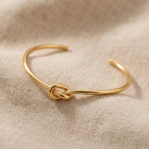 Puede incluir: Una pulsera de puño dorada con un diseño de nudo. La pulsera está hecha de una fina banda de metal curvada que forma un nudo en el centro. La pulsera está abierta en los extremos y parece estar hecha de metal brillante y pulido.