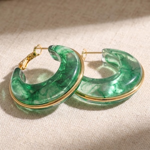 May include: A pair of hoop earrings featuring a translucent green marbled resin body with a polished gold-tone metal accent band running through the centre. The earrings include gold-tone metal post backings for secure attachment to the earlobe.