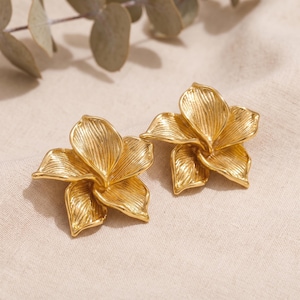 Op de afbeelding: Een paar goudkleurige metalen oorbellen in de vorm van open bloemen. Elke oorbel heeft vijf afzonderlijke, overlappende bloemblaadjes met een gestructureerd, lineair oppervlak dat vanuit het midden uitstraalt voor een gelaagd effect.