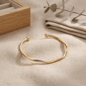 Könnte beinhalten: Ein goldfarbenes, offenes Armreif-Armband mit einem gedrehten Design. Das Armband hat eine glatte, polierte Oberfläche und ist mit kleinen, runden Kugelenden versehen. Der Schmuck wird auf einer neutralfarbenen Stoffoberfläche präsentiert.