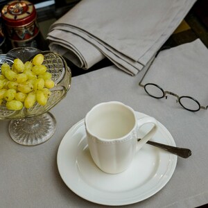 May include: A still life arrangement featuring a white teacup and saucer with a spoon, a glass bowl of green grapes, and a pair of round-framed glasses. A stack of folded gray napkins and decorative jars are also present.