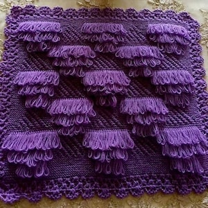 Puede incluir: Una manta cuadrada tejida de color morado con un patrón de puntos diagonales texturizados y varias filas de bucles de hilo unidos. Los bordes están rematados con un borde de ganchillo decorativo, creando una superficie uniforme y táctil.