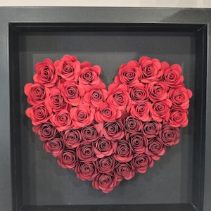Könnte beinhalten: Ein gerahmtes Herz aus roten Papierrosen. Die Rosen sind herzförmig angeordnet, mit einem Farbverlauf aus roten Farbtönen. Der Rahmen ist schwarz und der Hintergrund ist dunkelgrau. Dieses dekorative Stück eignet sich für die Inneneinrichtung.