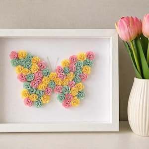 May include: A framed butterfly art piece made of colorful paper roses in pink, yellow, and green. The butterfly is centered in a white frame. A white vase with pink tulips sits to the right of the frame.