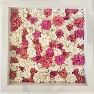 Könnte beinhalten: Gerahmtes Blumenarrangement mit einer dichten Ansammlung von Papierblumen. Die Blumen sind in Weiß-, Rosa- und Fuchsiatönen gehalten und bilden eine lebendige, strukturierte Darstellung. Der Rahmen ist weiß.