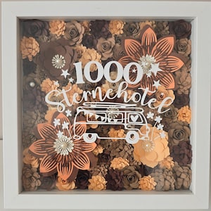 Peut inclure: Un cadre vitrine carré blanc rempli de fleurs en papier superposées dans des tons marron, beige et pêche. La vitre porte l'inscription blanche 1000 Sternehotel au-dessus d'un dessin au trait représentant un camping-car avec des étoiles.