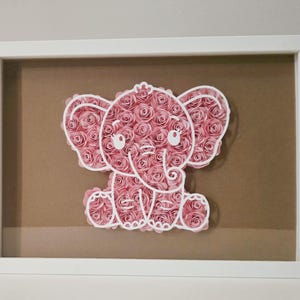 May include: A framed artwork featuring a pink elephant made of paper roses. The elephant is outlined in white and sits on a brown background within a white frame. The artwork is a decorative piece.