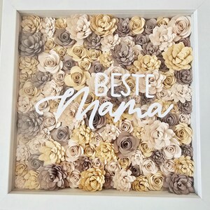 Könnte beinhalten: Gerahmtes Blumenarrangement mit weißem Rahmen. Die Anordnung zeigt zahlreiche Papierblumen in Creme-, Beige- und Grautönen. Die Worte "BESTE Mama" sind in weißer Schrift über die Mitte der Blumen geschrieben.
