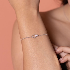 May include: A delicate silver bracelet with a small, circular clasp embellished with sparkling crystals. The fine chain of the bracelet is visible against the skin, creating a minimalist and elegant aesthetic. The jewelry is designed for everyday wear.