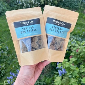 Puede incluir: Dos bolsas de papel marrón con golosinas para perros se sostienen frente a un fondo verde. Cada bolsa tiene una ventana transparente que muestra las golosinas. Las bolsas tienen una etiqueta azul que dice "Venison Dog Treats" y la marca "Hugo & Co."