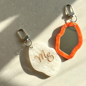 Può includere: Due portachiavi di forma irregolare con chiusure a moschettone in metallo color argento. Un portachiavi è bianco con una trama marmorizzata e presenta il testo in corsivo dorato Me. L'altro ha un bordo marmorizzato arancione brillante.