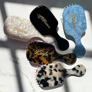 May include: Five hairbrushes with personalized gold lettering. One is white pearlescent with 'Marissa', one is solid black with 'Allison', one is blue marbled with 'Starver', one is brown tortoiseshell with 'Kayla', and one is white with black spots with 'Mrs. Harper'.