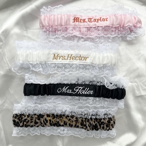 May include: Four elasticized garters with white lace trim. The top is pink satin with 'Mrs. Taylor' embroidered in brown. The second is white satin with 'Mrs. Hector' in gold. The third is black satin with 'Mrs. Holler' in white. The bottom is leopard print.