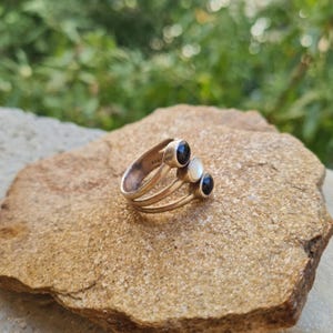 Pode incluir: Um anel de prata com três bandas distintas que convergem numa única haste. O anel é adornado com três pedras redondas em cabochão: duas pedras pretas nas extremidades e uma pedra branca no centro, todas cravadas em prata polida.