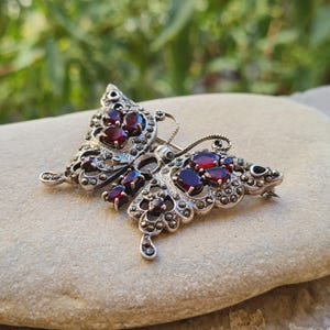 Può includere: Una spilla a forma di farfalla in argento con pietre granato rosso scuro dal taglio ovale incastonate nelle ali. La struttura in metallo è decorata con piccole pietre di marcasite grigio scuro disposte in motivi complessi sul corpo e sui bordi.