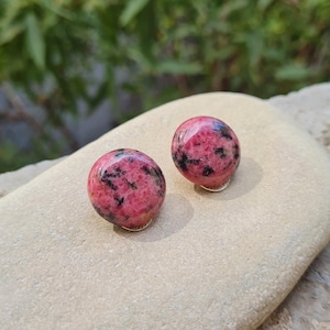 May include: A pair of round stud earrings featuring polished rhodonite stones with a vibrant pink base colour and distinct black mineral inclusions. The stones have a smooth, domed surface and are set on simple metal posts for secure attachment.