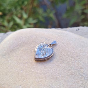May include: A silver-toned heart-shaped locket pendant featuring an intricate engraved scrollwork pattern on its front surface. The metal locket has a small triangular bail at the top for attachment to a chain, showcasing a polished metallic finish.