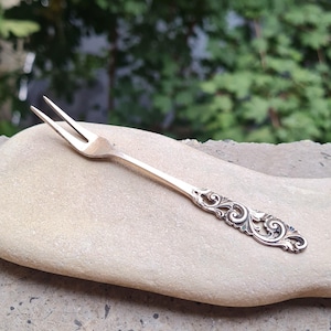 May include: A silver-toned metal cocktail fork featuring two slender tines and a decorative handle with intricate, raised scrollwork patterns. The handle displays a polished finish with darkened recessed areas that highlight the detailed relief design.