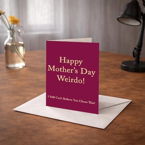 Op de afbeelding: Een bordeauxrode Moederdagkaart met de tekst "Happy Mother's Day Weirdo!" in een schreeflettertype. De kaart ligt op een witte envelop, met de tekst "I Still Can't Believe You Chose This!" onderaan.