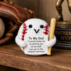 May include: A crocheted baseball figure with a smiling face and a small wooden bat. The baseball holds a sign that reads "To My Dad, I scored a home run by getting you as my dad! love you to the ballpark and back."