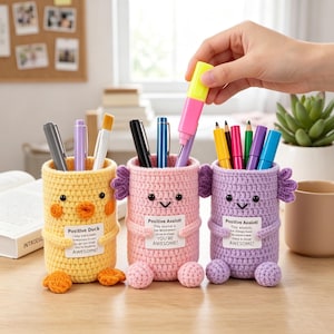 May include: Three cylindrical crochet pen holders shaped like animals. The yellow one is a duck, while the pink and purple ones are axolotls. Each features a small white label with text, black bead eyes, and small crocheted limbs and feet.