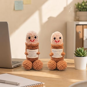 May include: Two handmade crochet penis dolls. One doll wears glasses. Each doll has a smiling face and a sign that reads "Positive J" and "Positive Penis". The dolls are brown, tan, and peach coloured.