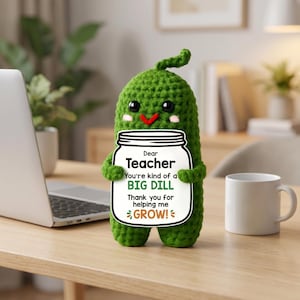 Op de afbeelding: Een handgemaakte groene gehaakte augurkenfiguur met een bord met de tekst "Dear Teacher, You're kind of a BIG DILL, Thank you for helping me GROW!". De augurk heeft zwarte ogen en een rode glimlach. Een witte mok en laptop staan op het houten bureau.