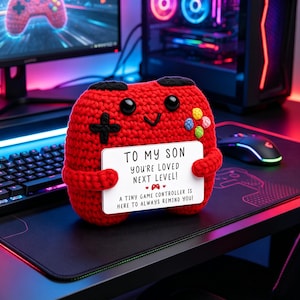 May include: A red crocheted game controller with a smiling face and black accents. It holds a white sign that reads "TO MY SON, YOU'RE LOVED NEXT LEVEL! A TINY GAME CONTROLLER IS HERE TO ALWAYS REMIND YOU!" A fun gift for gamers.