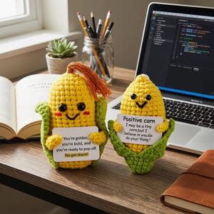 May include: Two handmade crochet corn figures with smiling faces and holding signs with positive messages. One says "You're golden, you're bold, and you're ready to pop off. Go get them!" The other says "Positive corn I may be a tiny corn, but I believe in you. Go do your thing!"