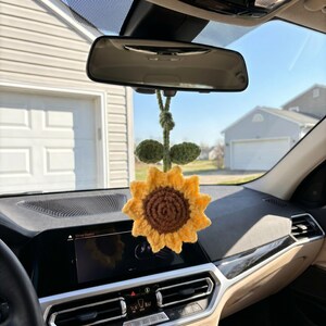 May include: A crocheted sunflower car charm with yellow petals and a brown center, hanging from a car's rearview mirror. The charm includes green leaves and a stem, adding a touch of nature to the car's interior.
