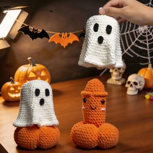 May include: Crocheted Halloween decorations featuring a ghost and a pumpkin-shaped figure. The ghost is white with black eyes and mouth, and the pumpkin figure is orange with a smiling face. The background includes pumpkins, skulls, and a bat garland.