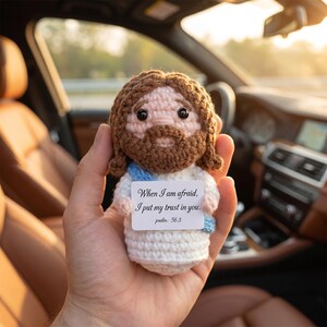 May include: A handmade crochet doll of Jesus with brown hair and beard, wearing a white robe and blue sash. A small card with the text "When I am afraid, I put my trust in you. psalm: 56:3" is attached. The doll is held in a hand.