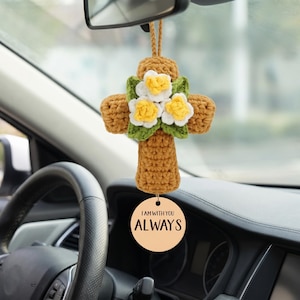 Può includere: Un ciondolo a forma di croce all'uncinetto, nei toni del marrone, verde, bianco e giallo, appeso a uno specchietto retrovisore. La croce è decorata con fiori e un'etichetta di legno con la scritta "I AM WITH YOU ALWAYS".